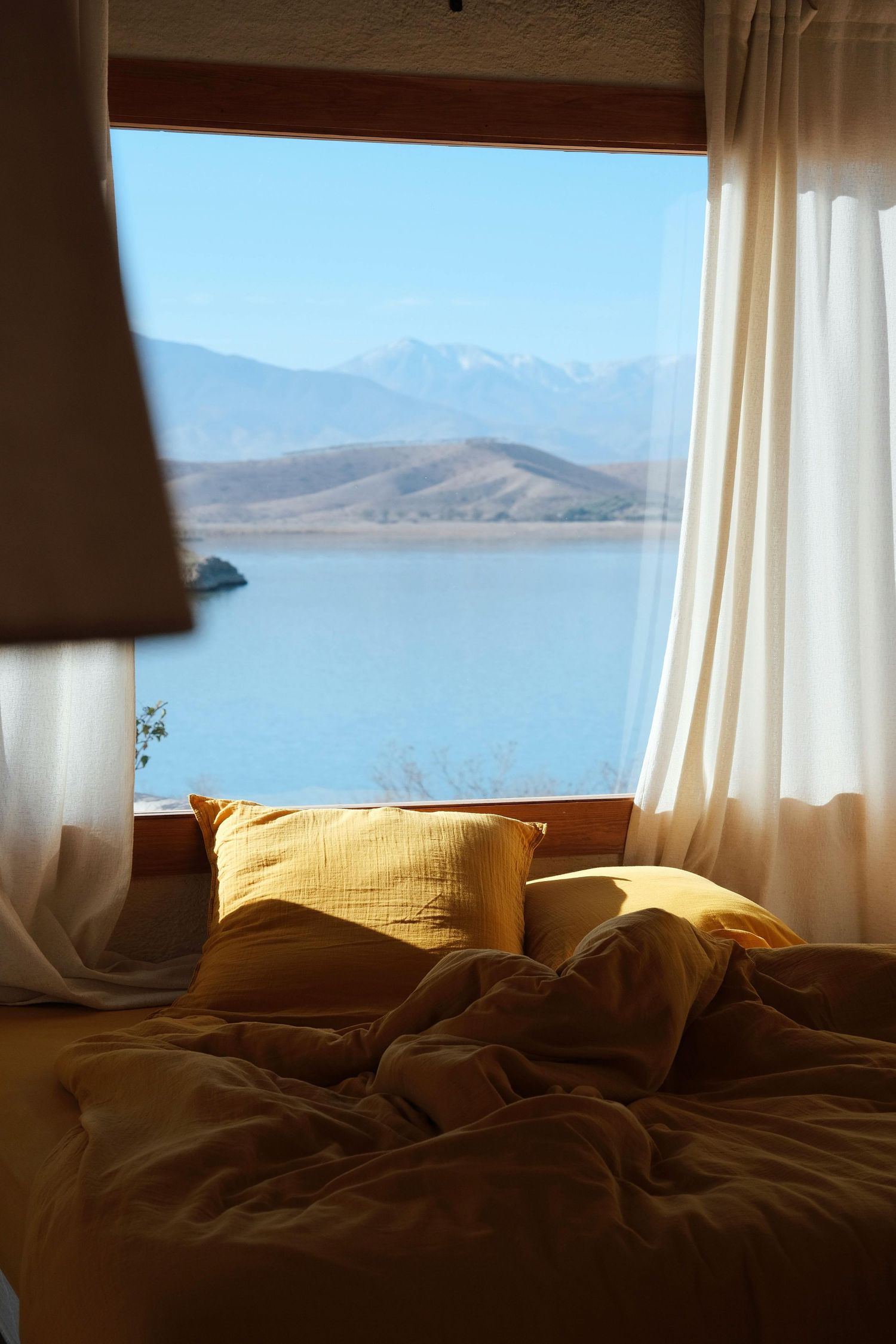 Cozy unmade bed with yellow pillows by window overlooking serene mountain lake and distant peaks on sunny day.