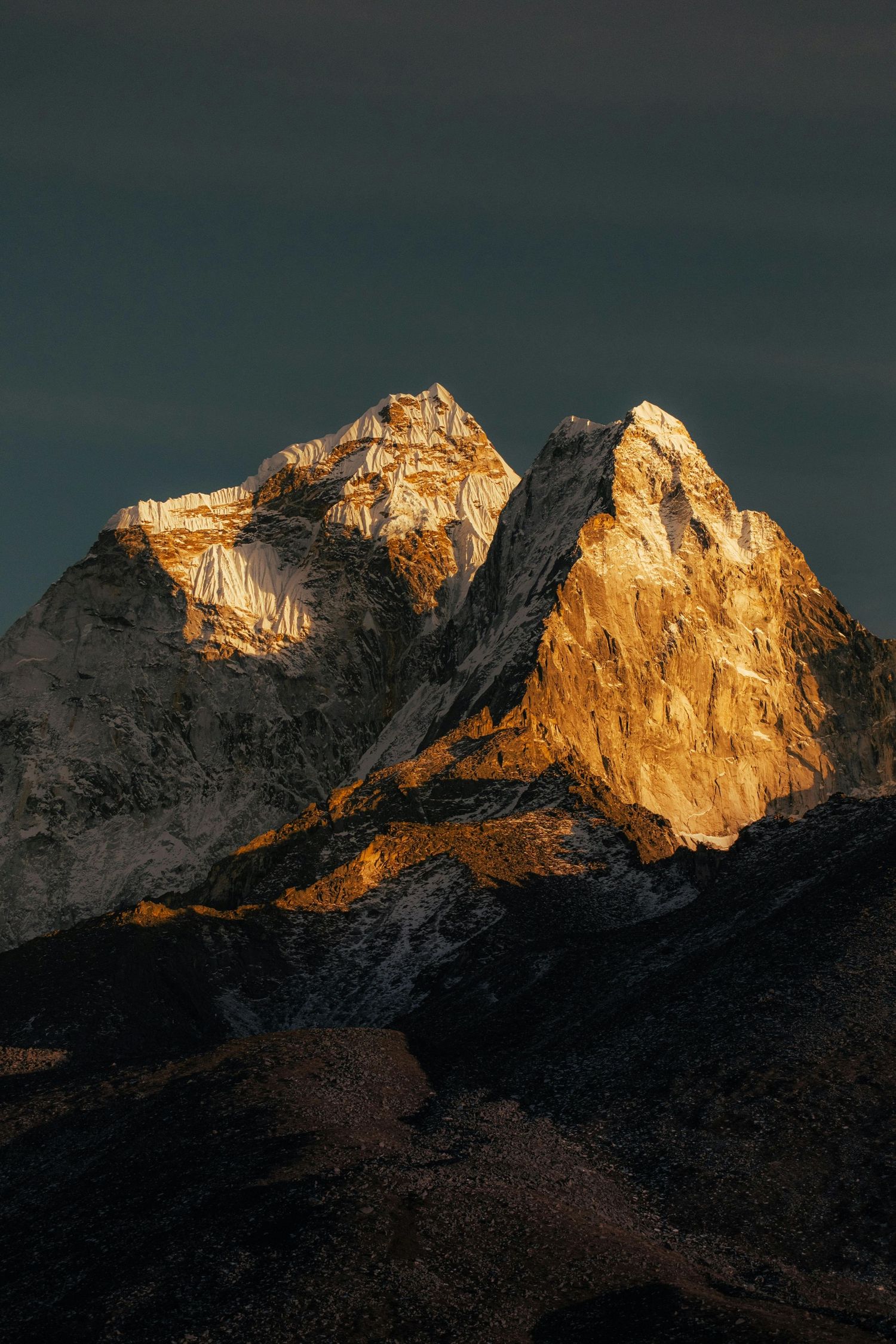 Snow-capped mountain peaks catch golden sunlight against a dark moody sky at sunset.