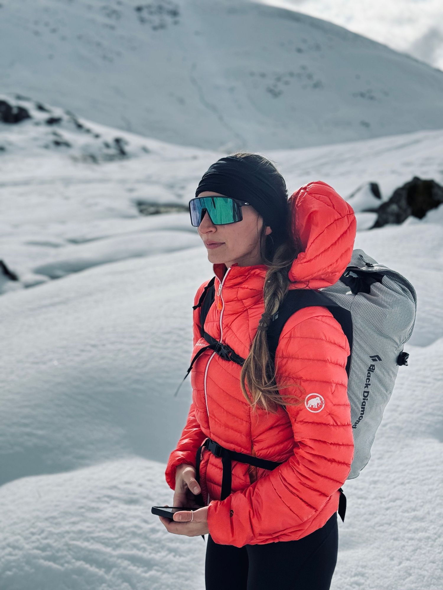 A winter sports enthusiast in a bright orange puffer jacket carries a backpack while navigating snowy mountain terrain.
