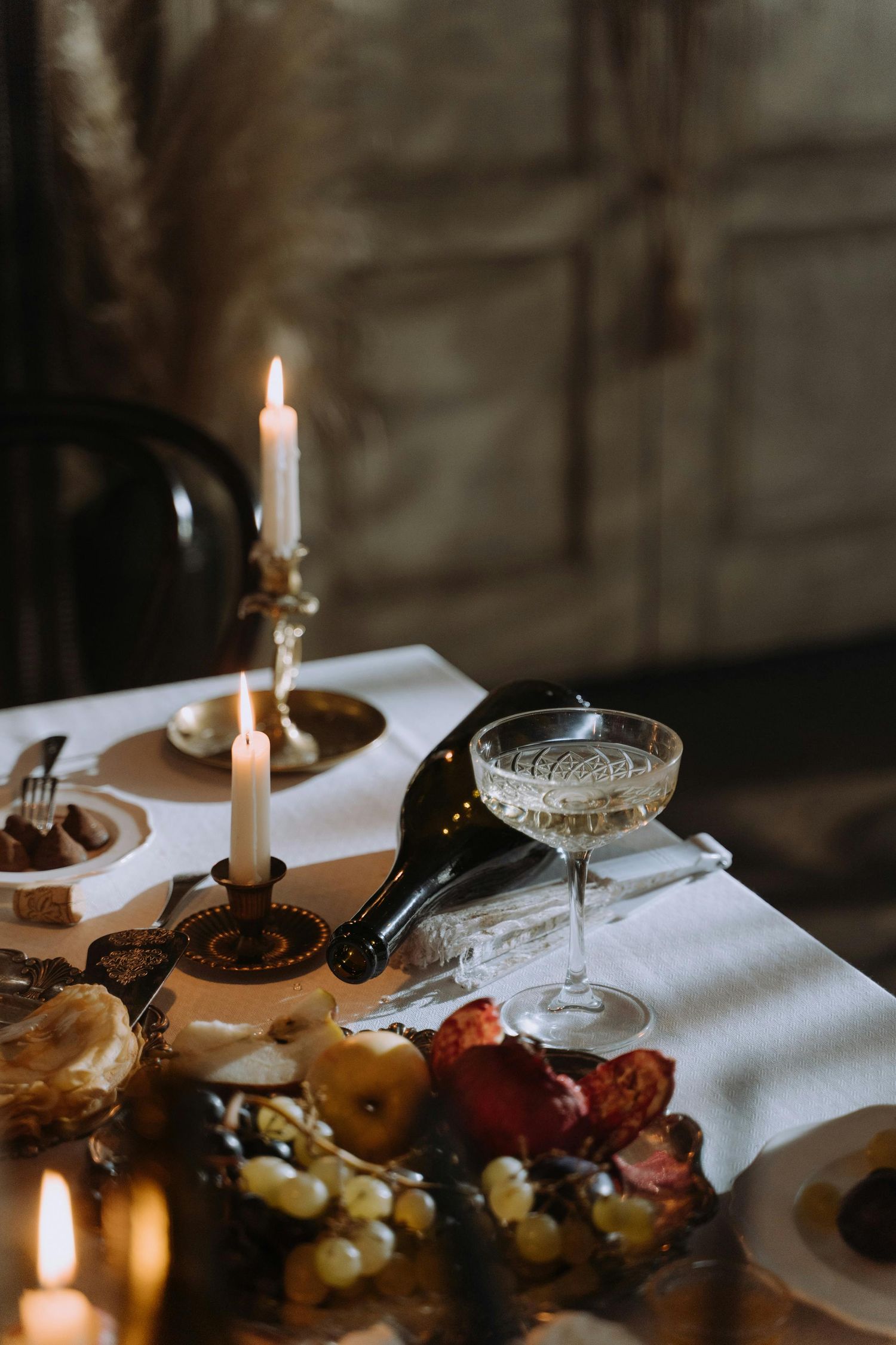Elegant table setting with lit candles, coupe champagne glass, and fresh fruit on white linen tablecloth in moody lighting.