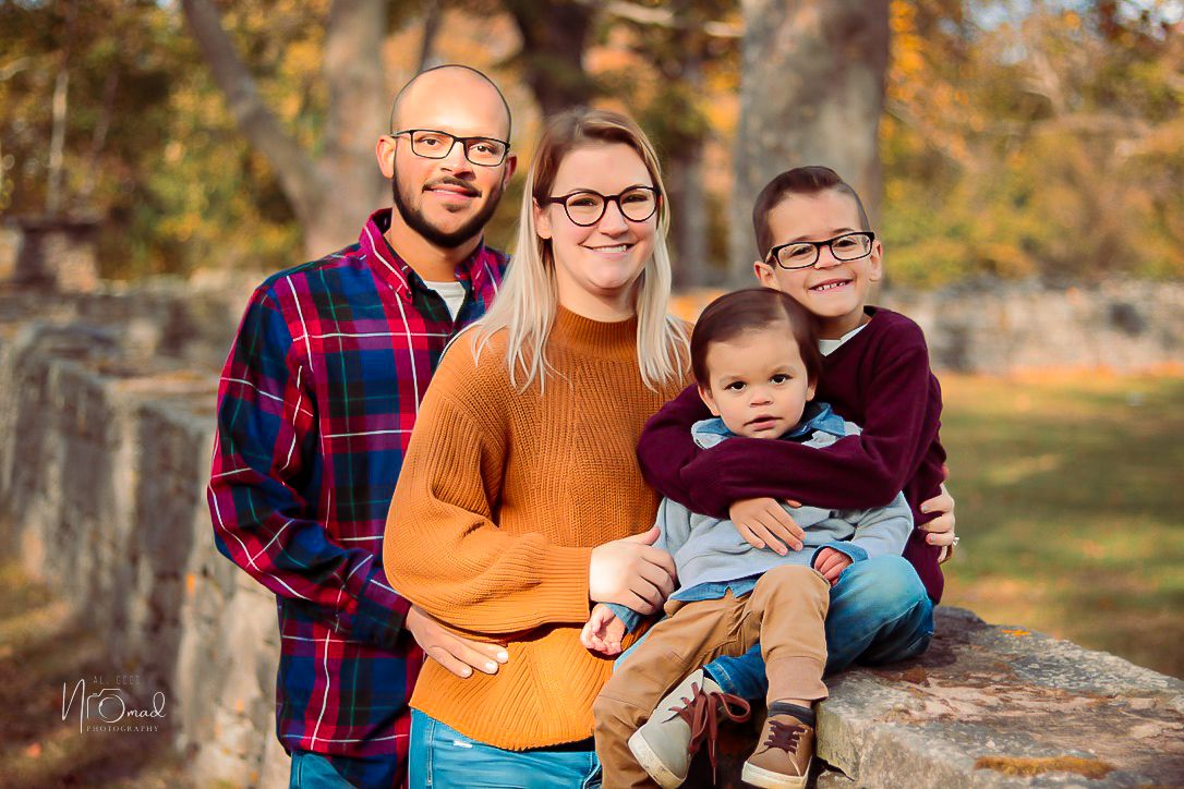 A family wearing fall-colored clothing poses together outdoors against autumn foliage and stone wall backdrop.