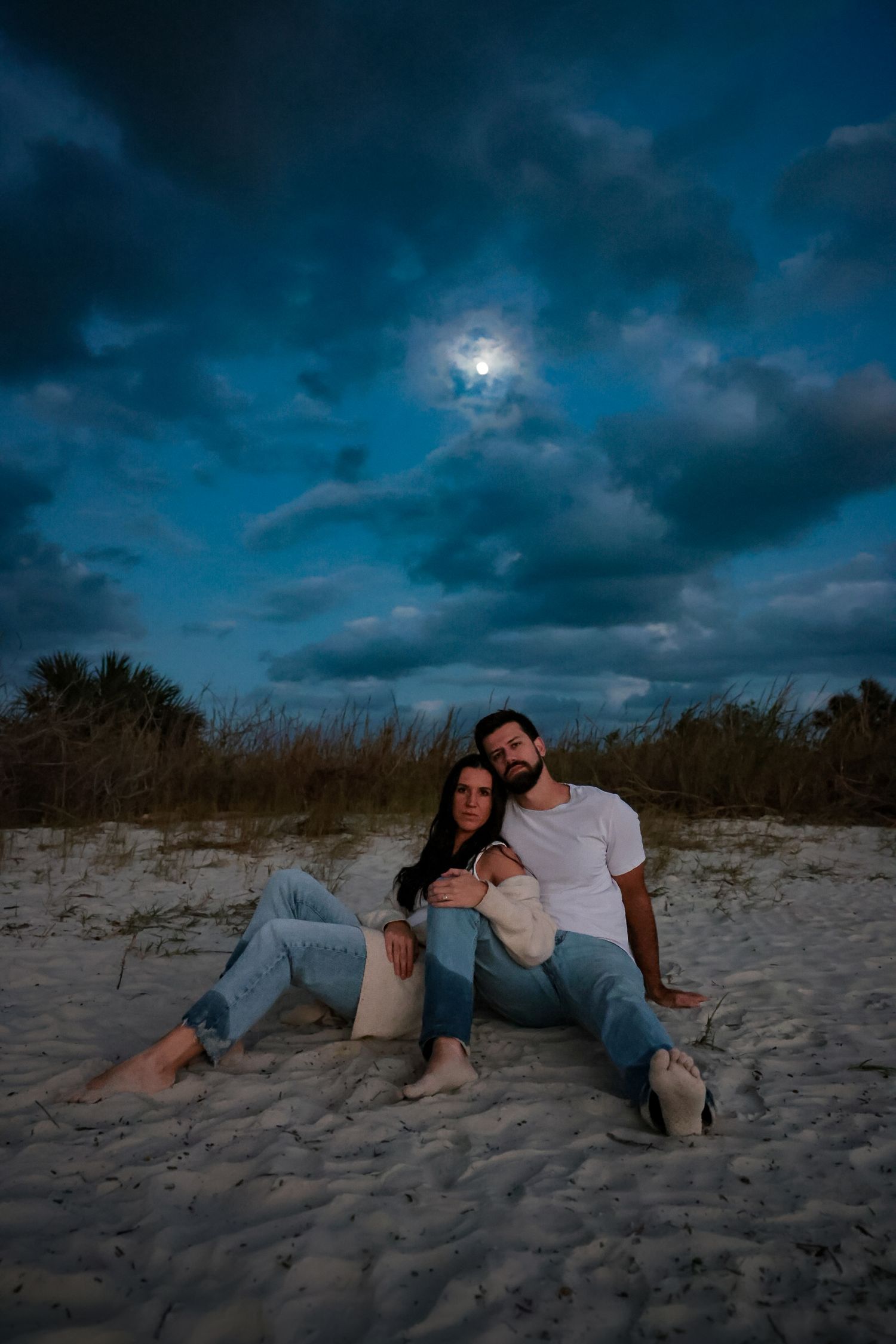 A couple sits together on a sandy beach at dusk with dramatic dark clouds and moonlight illuminating the evening sky.
