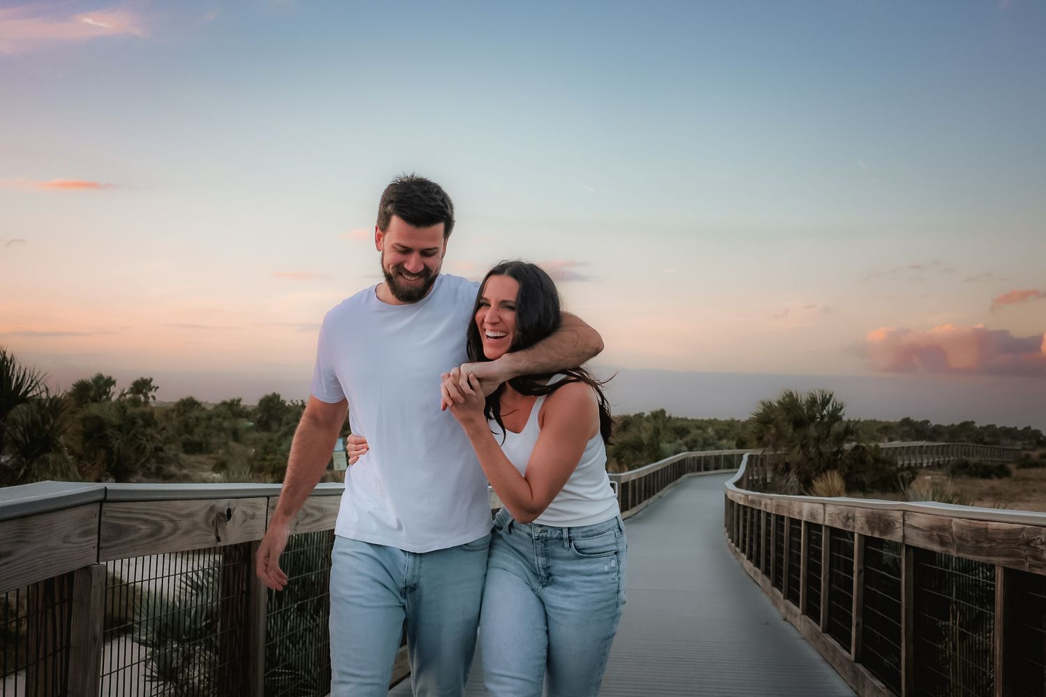 A happy couple embraces on a scenic boardwalk at sunset with ocean views and palm trees in the background.