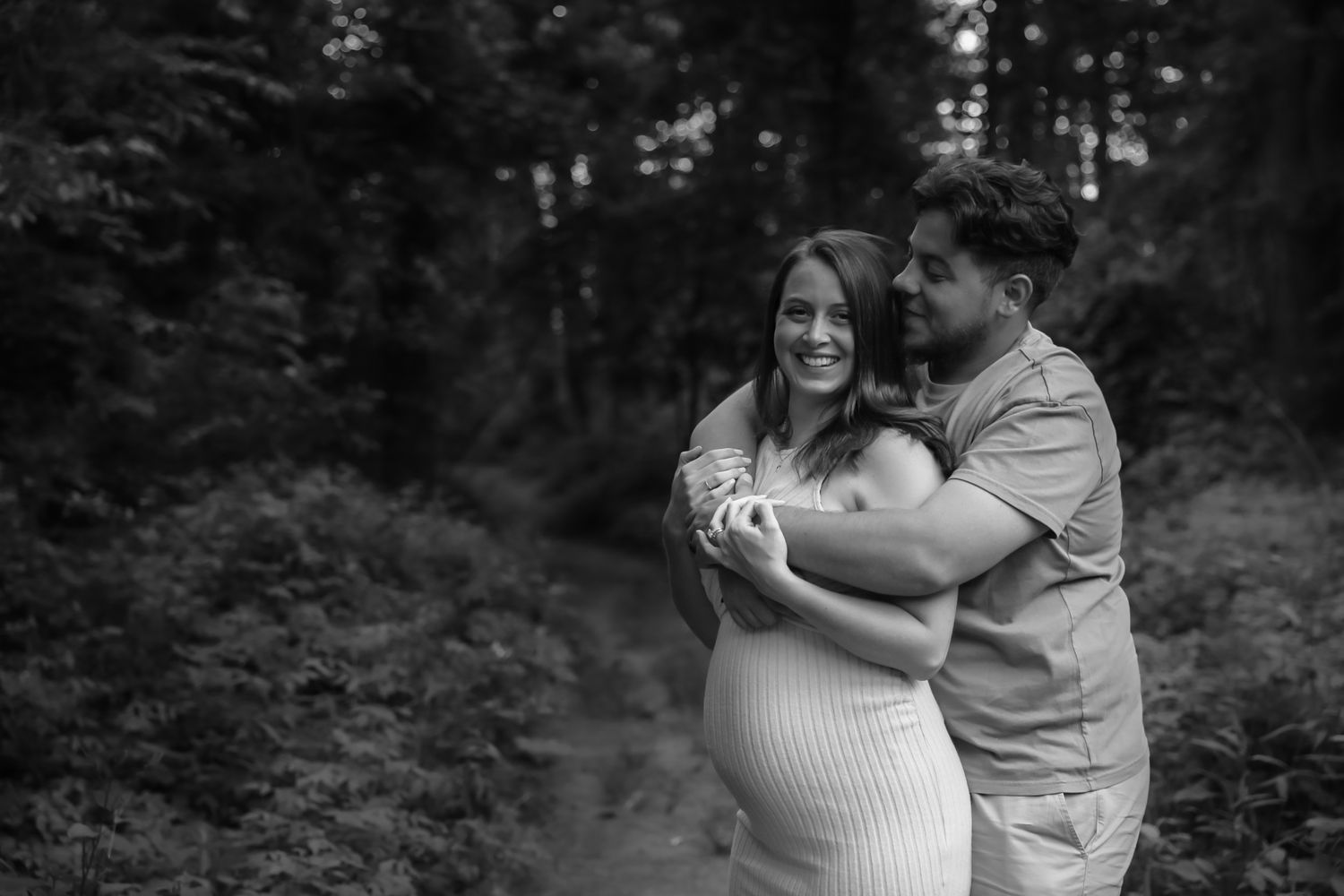 Black and white maternity photo of a smiling couple embracing outdoors in a lush forest setting.