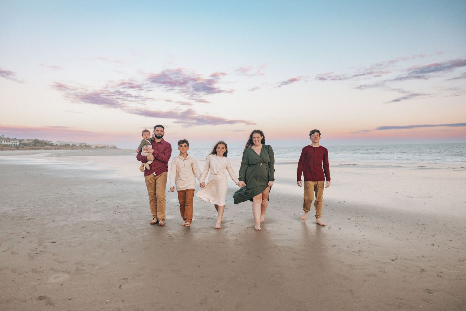 Family of six walking on a beach at sunset, wearing coordinated outfits in warm tones, with ocean waves in the background.