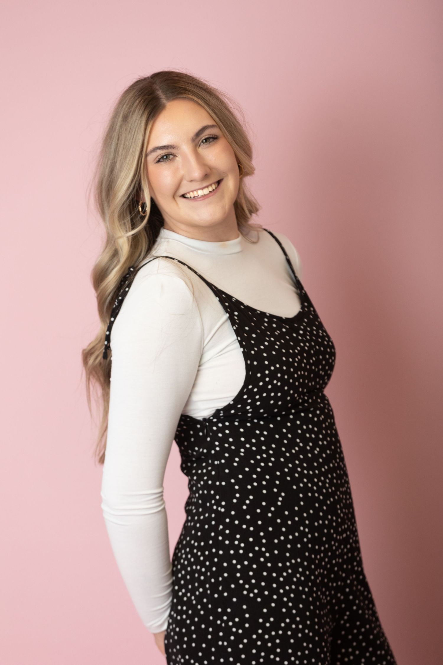 Studio portrait in polka dot dress and white turtleneck against pink background.