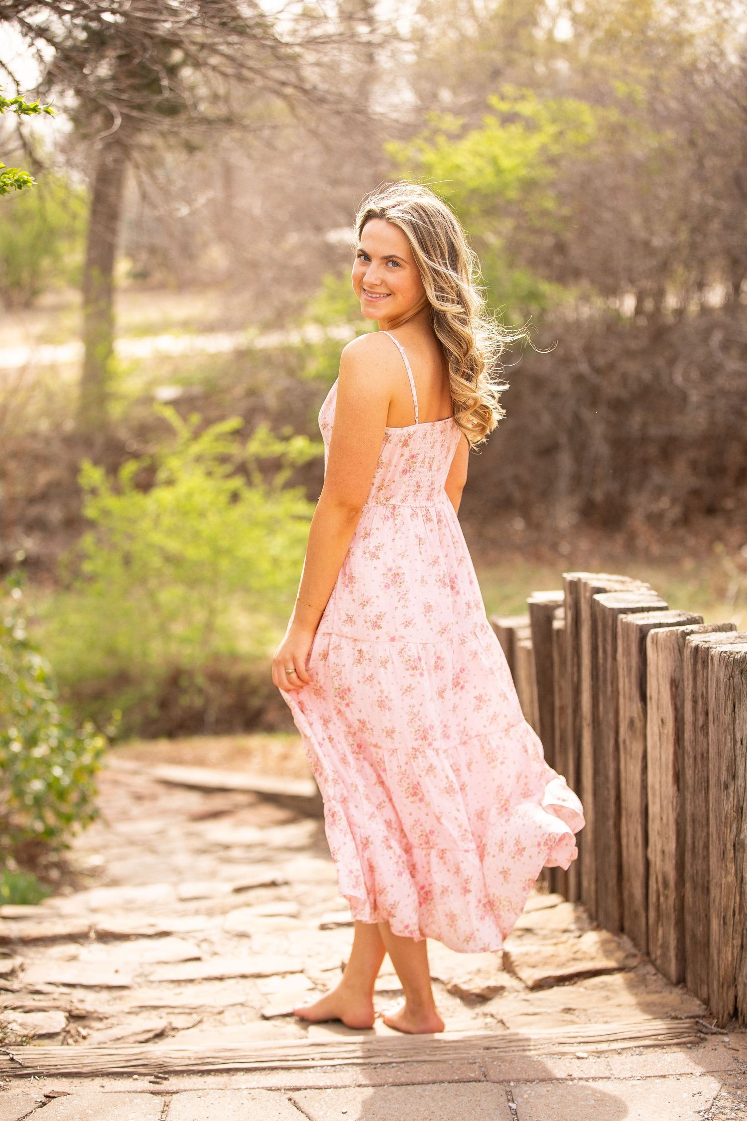 A woman in a flowing pink floral dress poses gracefully on a wooden walkway in a sunlit outdoor setting.
