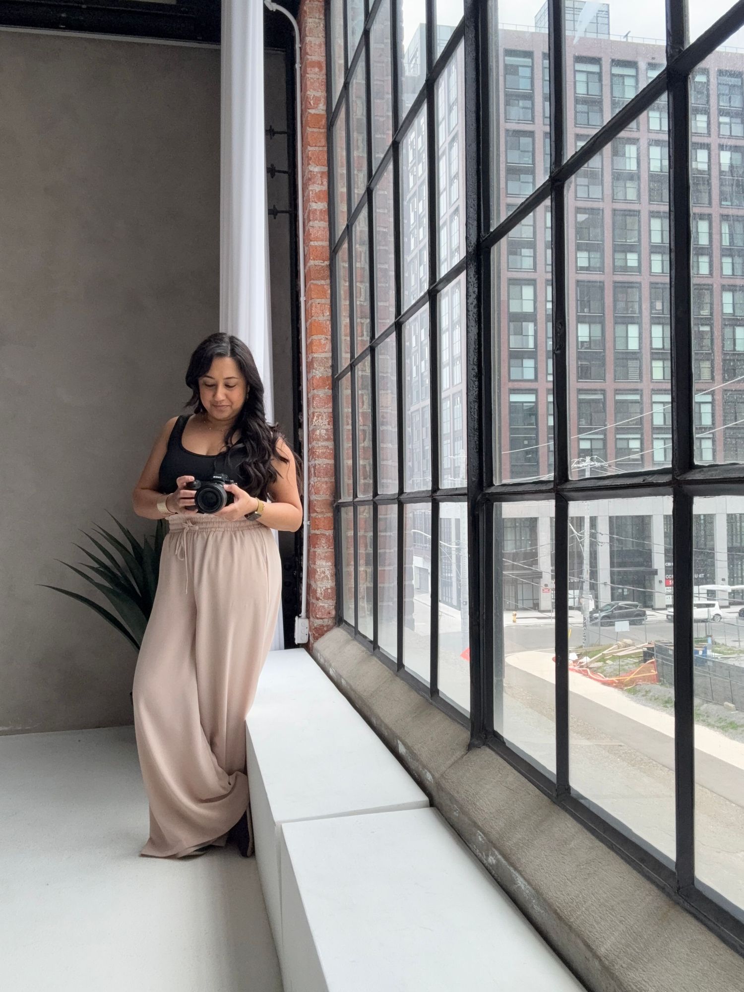 Woman in beige maxi dress holding camera stands by large industrial windows with city building views.