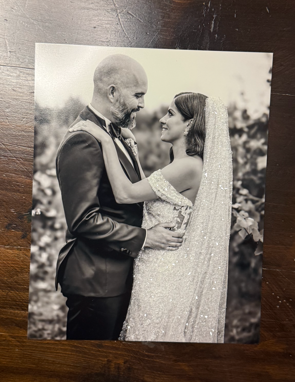 Black and white wedding photo of a smiling bride in a sparkly gown and groom in a tuxedo embracing outdoors.