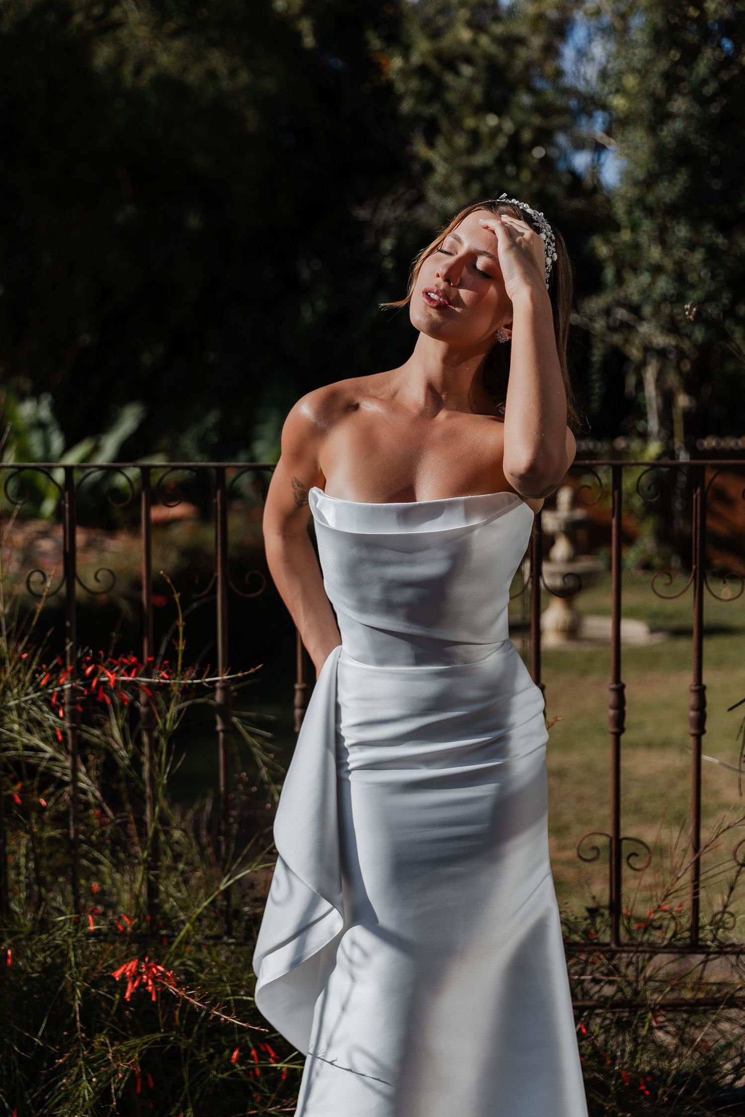 A figure in a white strapless dress poses gracefully against an outdoor garden background with iron fencing.