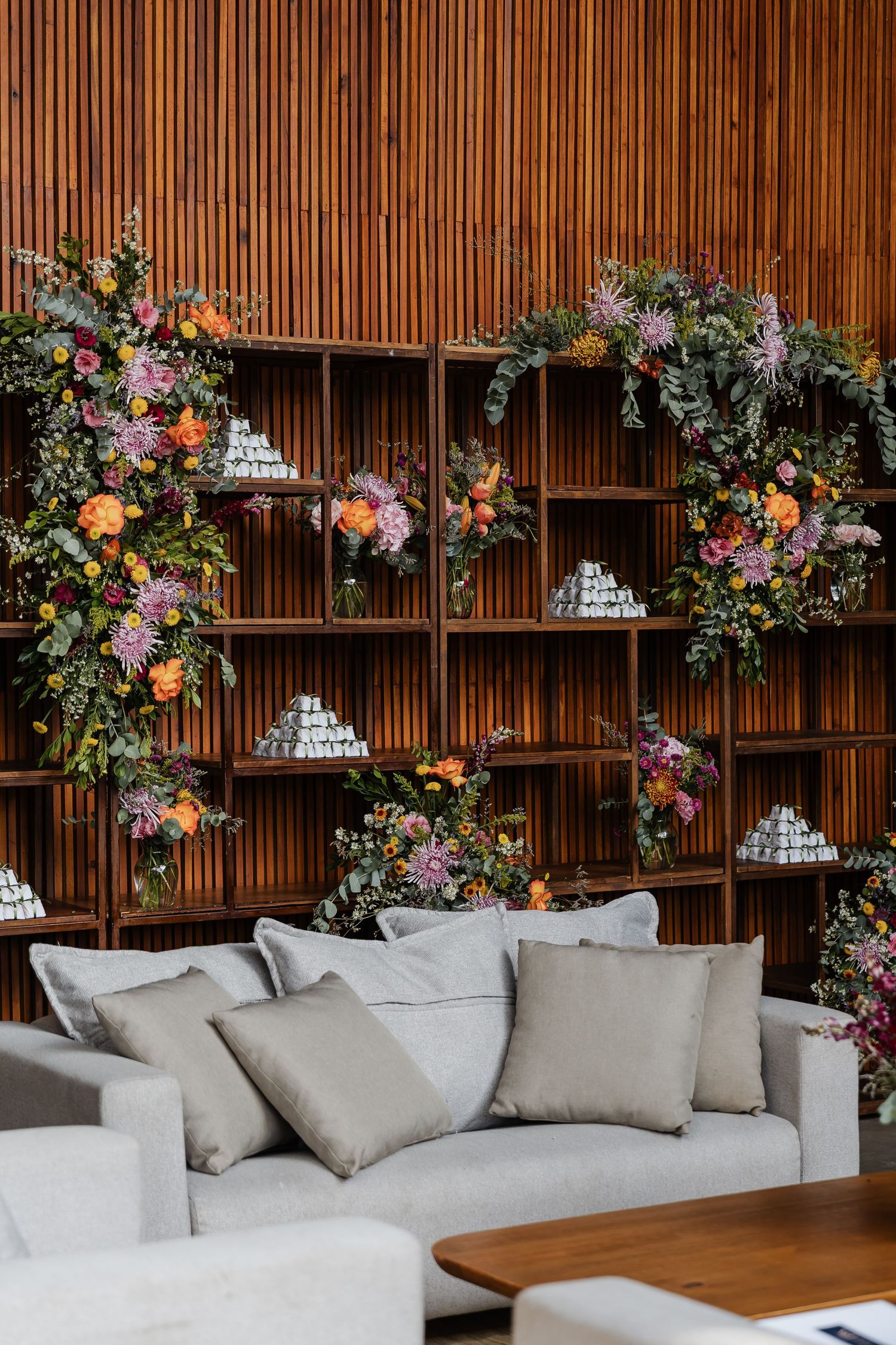 casamento em Belo HorizoModern wood-paneled lounge area with white sectional sofa and colorful floral arrangements mounted on wall shelving.