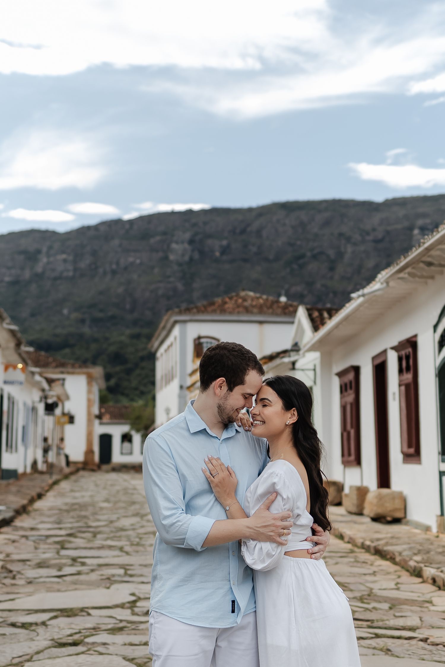 A couple embraces on a cobblestone street with white colonial buildings and mountains in the background in Brazil.