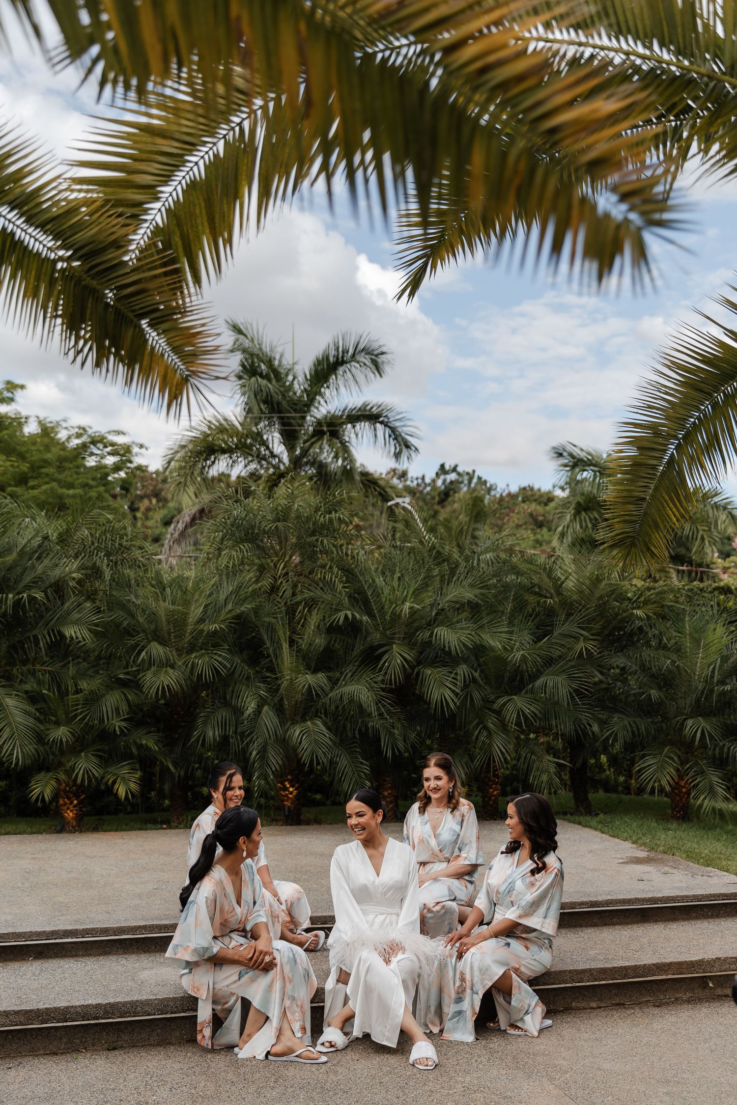 A group in white robes sits together on stone steps surrounded by lush tropical palm trees on a sunny day.