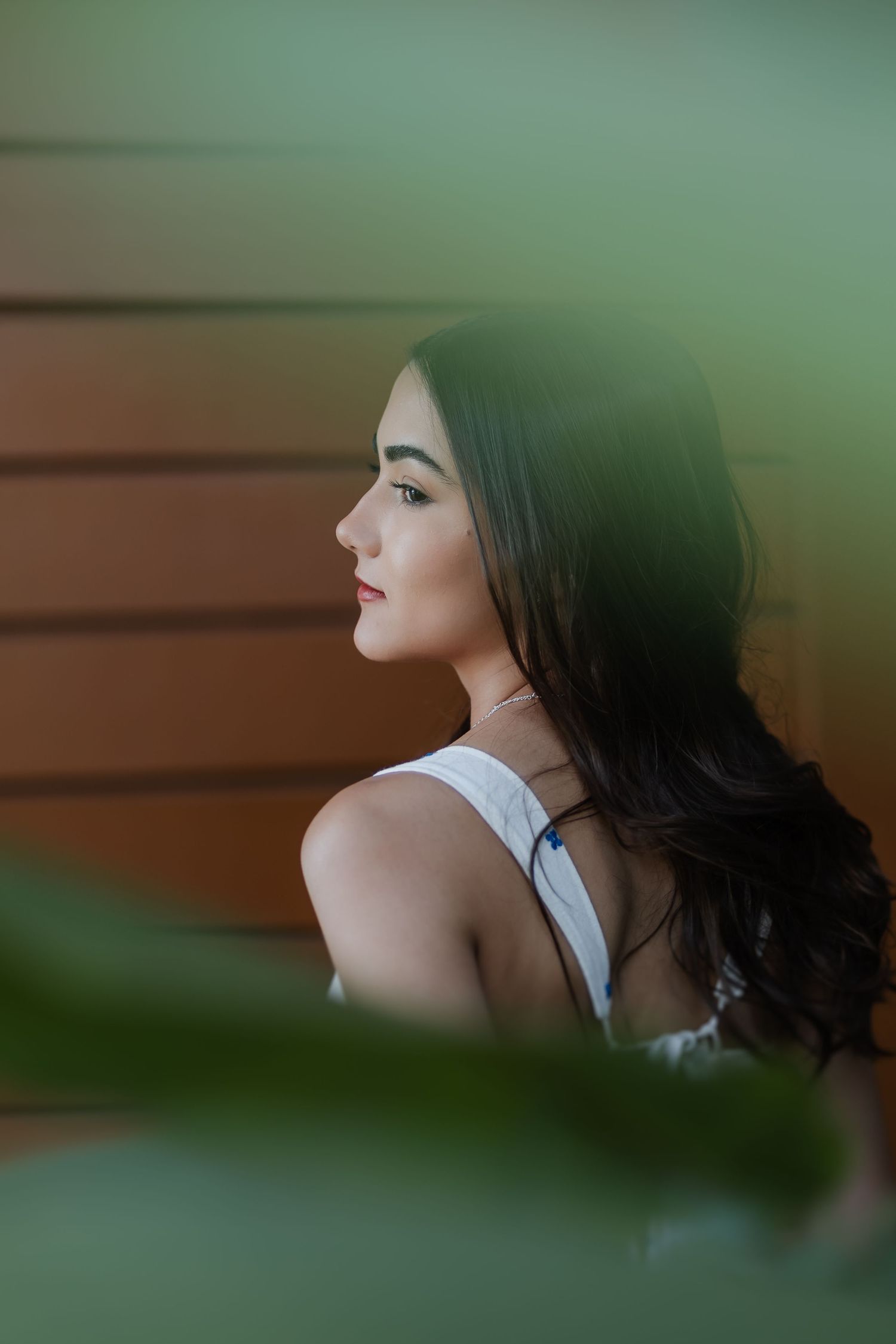 Side profile portrait through green leaves showing long dark hair and white sleeveless top against brown wall background.