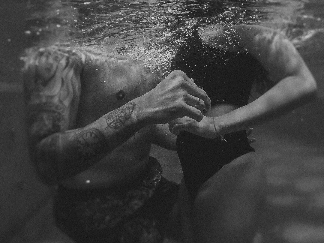Two people's hands clasped together underwater in an artistic black and white photograph showing air bubbles and movement.