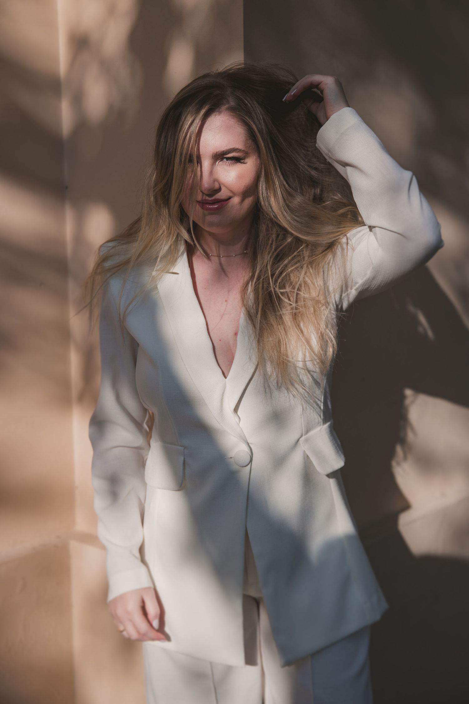 Person in white blazer and suit with flowing blonde hair poses against a wall with dramatic shadows and lighting.