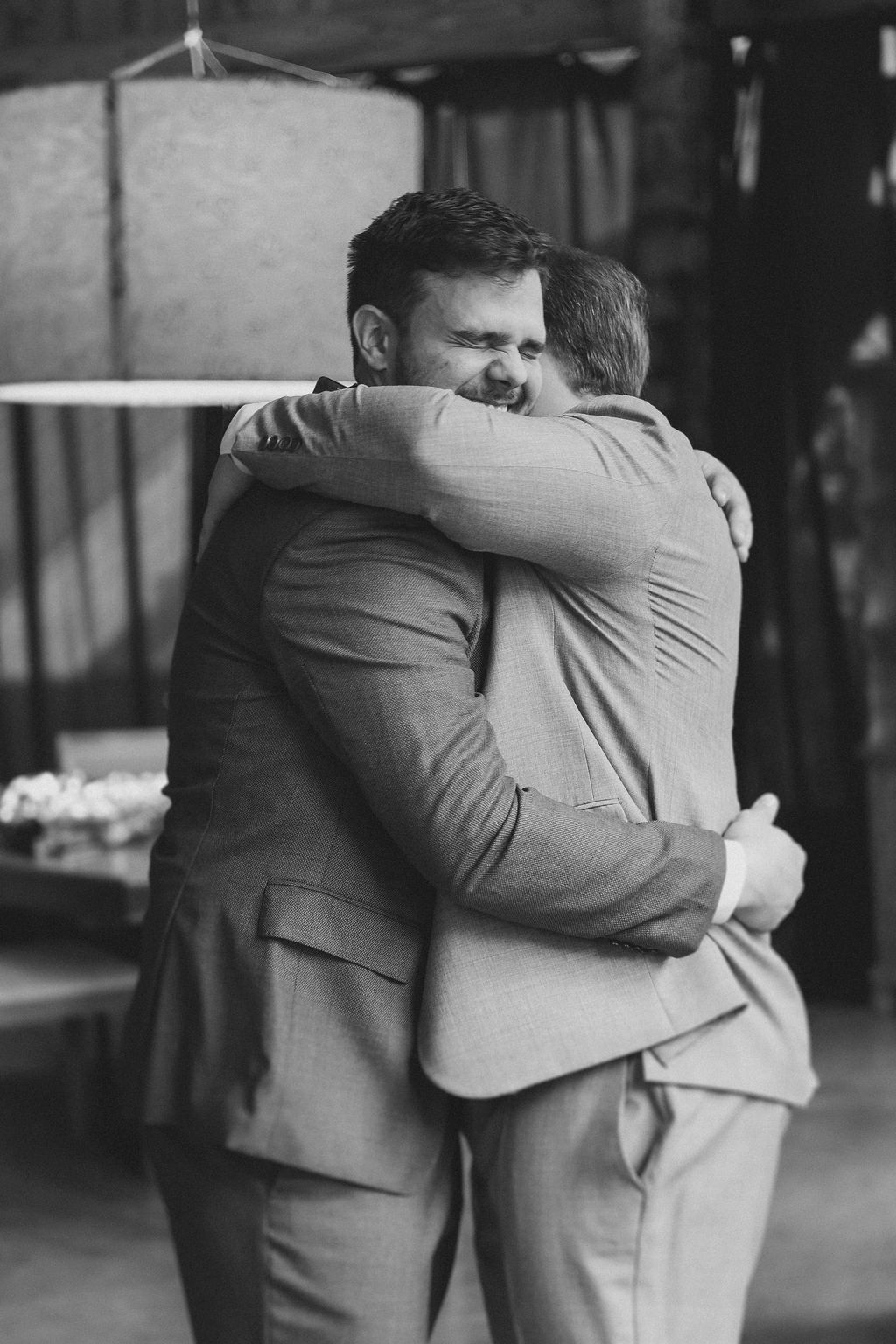 Inspire-se com o casamento ao ar livre eTwo people in light-colored suits share an emotional embrace in a candid black and white photograph.
