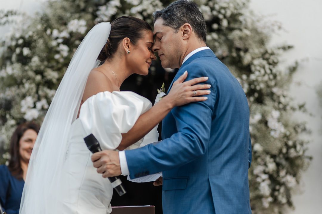 A tender wedding ceremony moment between a bride in white and groom in blue during their outdoor celebration.