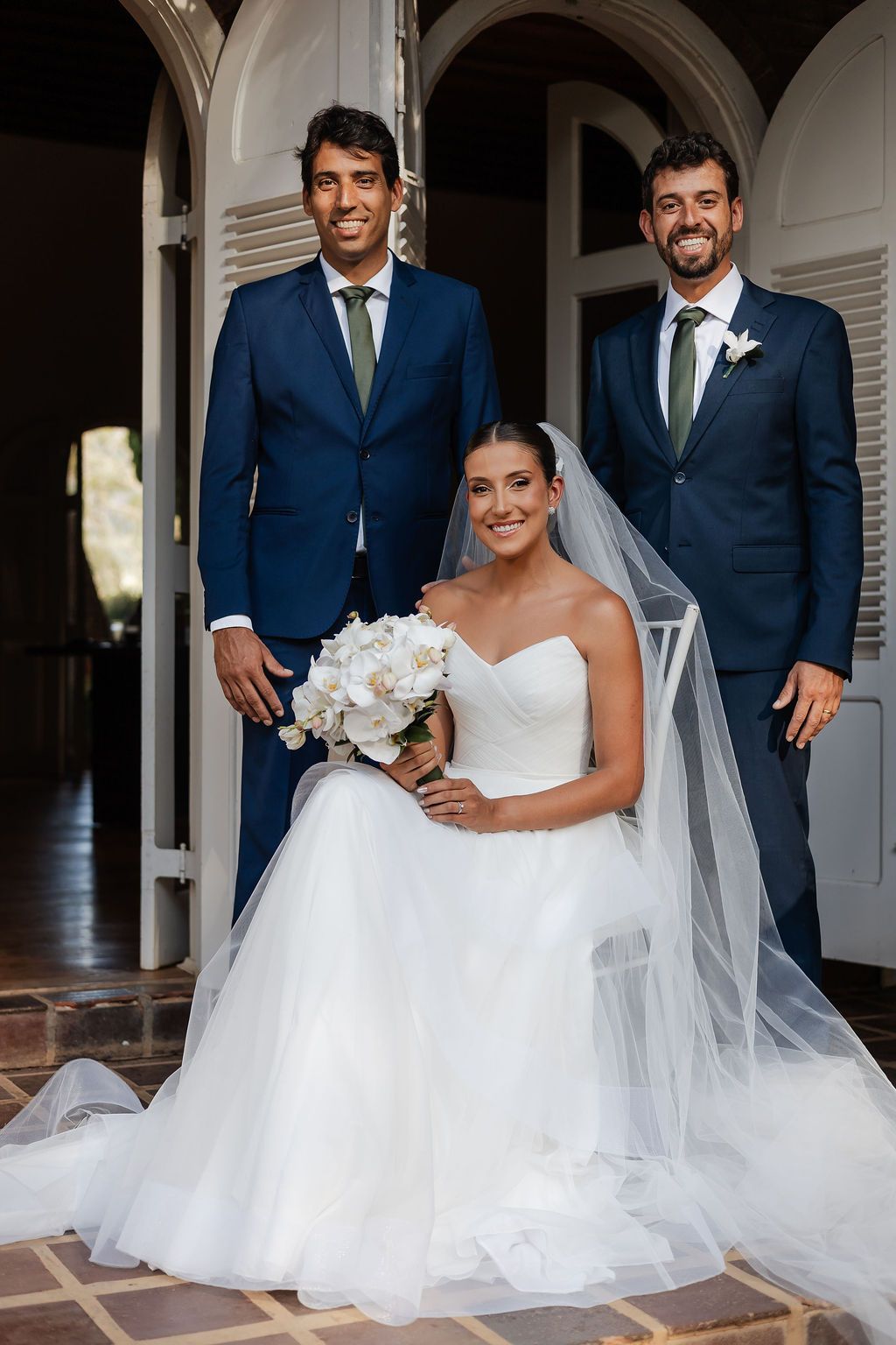 Cerimônia de casamento ao ar livre na FazeA bride in a white strapless gown with a flowing veil poses with two groomsmen in navy blue suits.