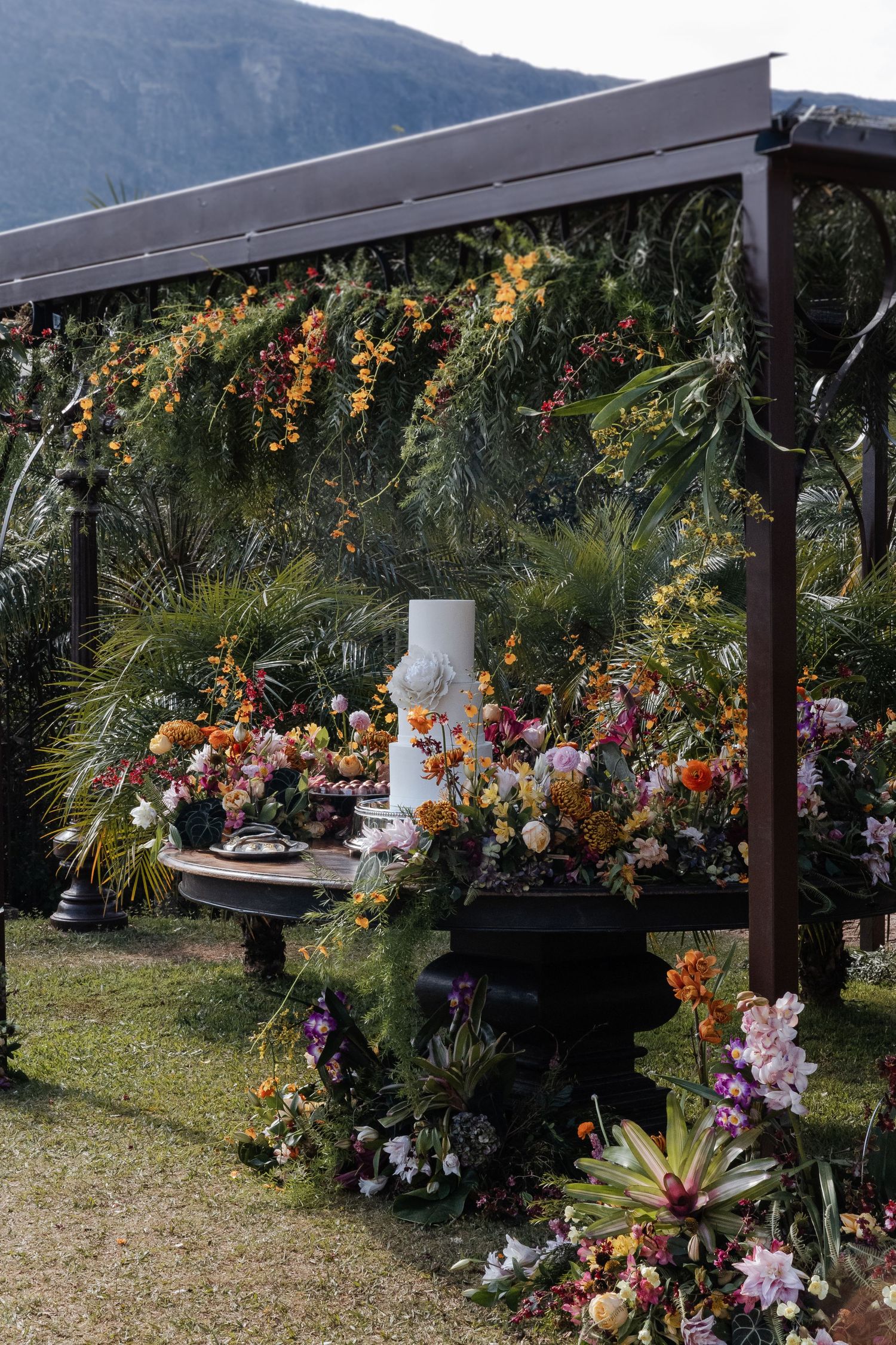 A rustic wooden pergola decorated with vibrant floral arrangements against a mountainous backdrop in a garden setting.