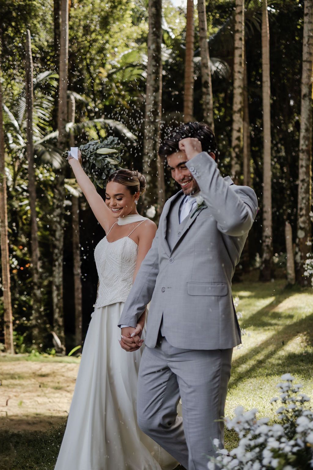 Joyful newlyweds celebrate together in a sunlit forest setting with flowers and tall trees.