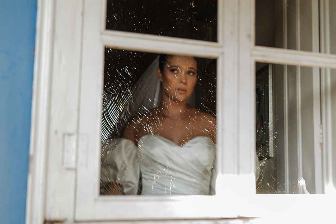Through a frosty window, someone in a white wedding dress gazes outward with long curly hair falling past their shoulders.