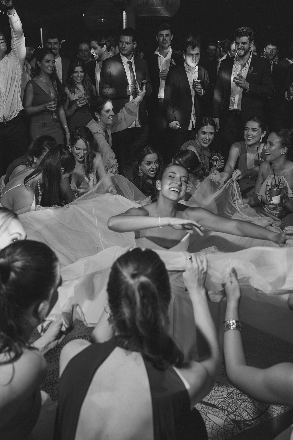 A group of partygoers celebrate as a figure lying on white sheets at a festive gathering is surrounded by revelers.