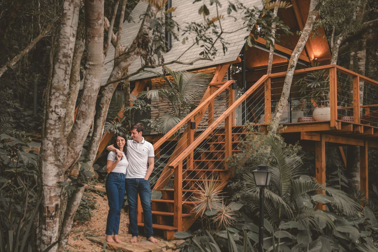 A couple stands together in a wooded setting next to a rustic staircase leading to a treehouse deck at dusk.
