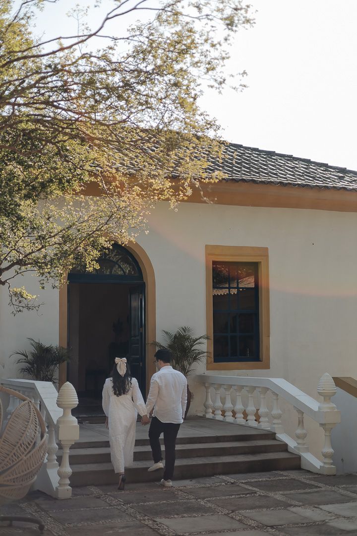 A couple walking up the steps to an elegant cream-colored house with arched doorway and decorative balustrades.