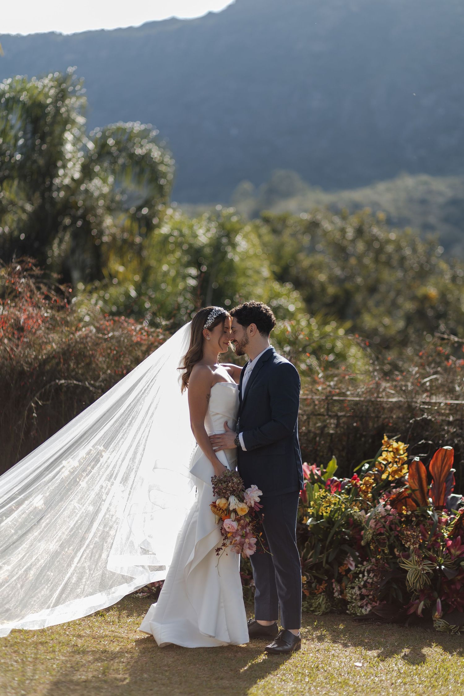 A long white wedding veil flows behind a couple embracing in a mountainside garden with colorful flowers.