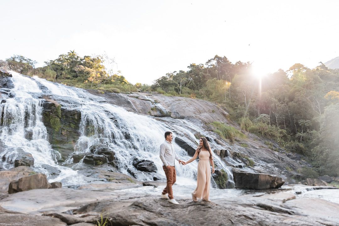 A couple walks hand in hand on a rocky waterfall during golden hour as sunlight streams through the trees.