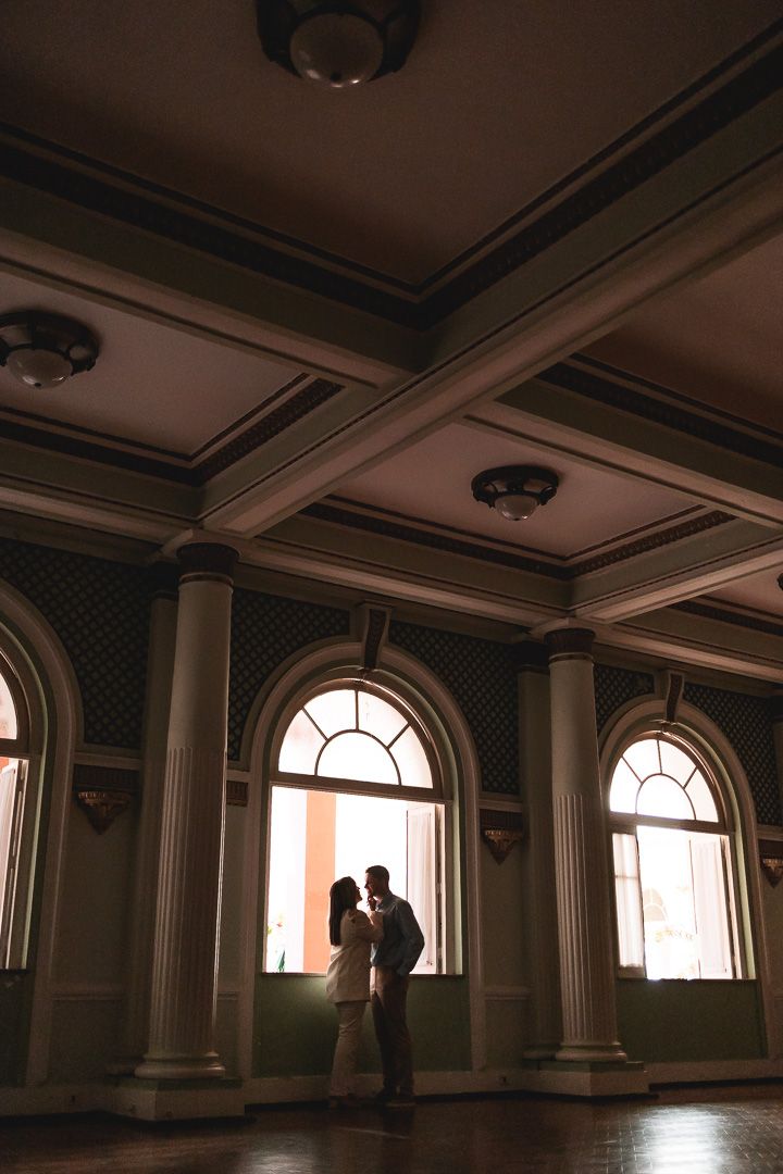 A silhouetted couple stands in an elegant ballroom with arched windows, columns and ornate ceiling details.