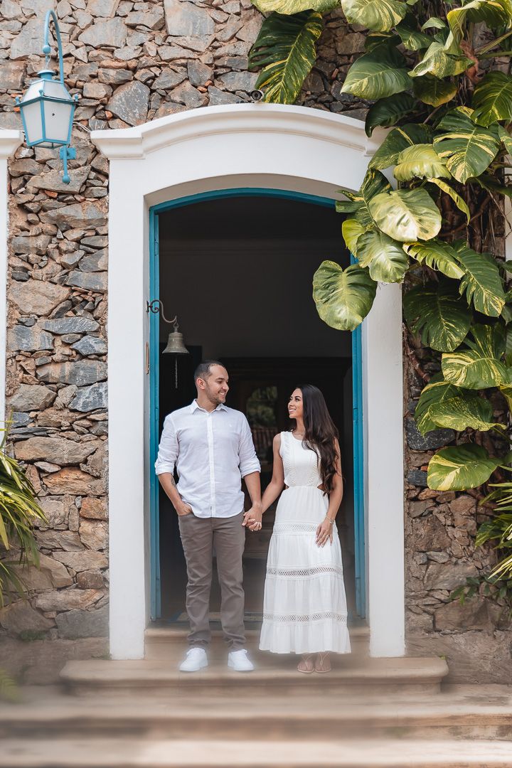 A couple stands in an ornate doorway with blue trim against a stone wall adorned with tropical monstera leaves and a lantern.