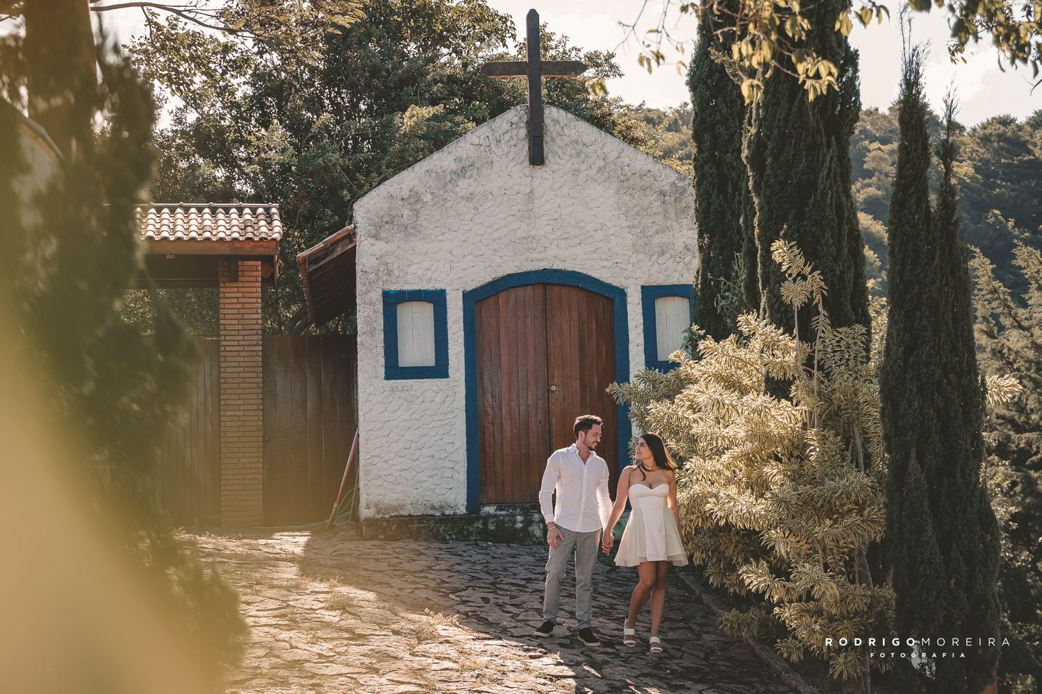 A couple strolls in front of a small white chapel with blue trim and wooden doors in a garden setting.