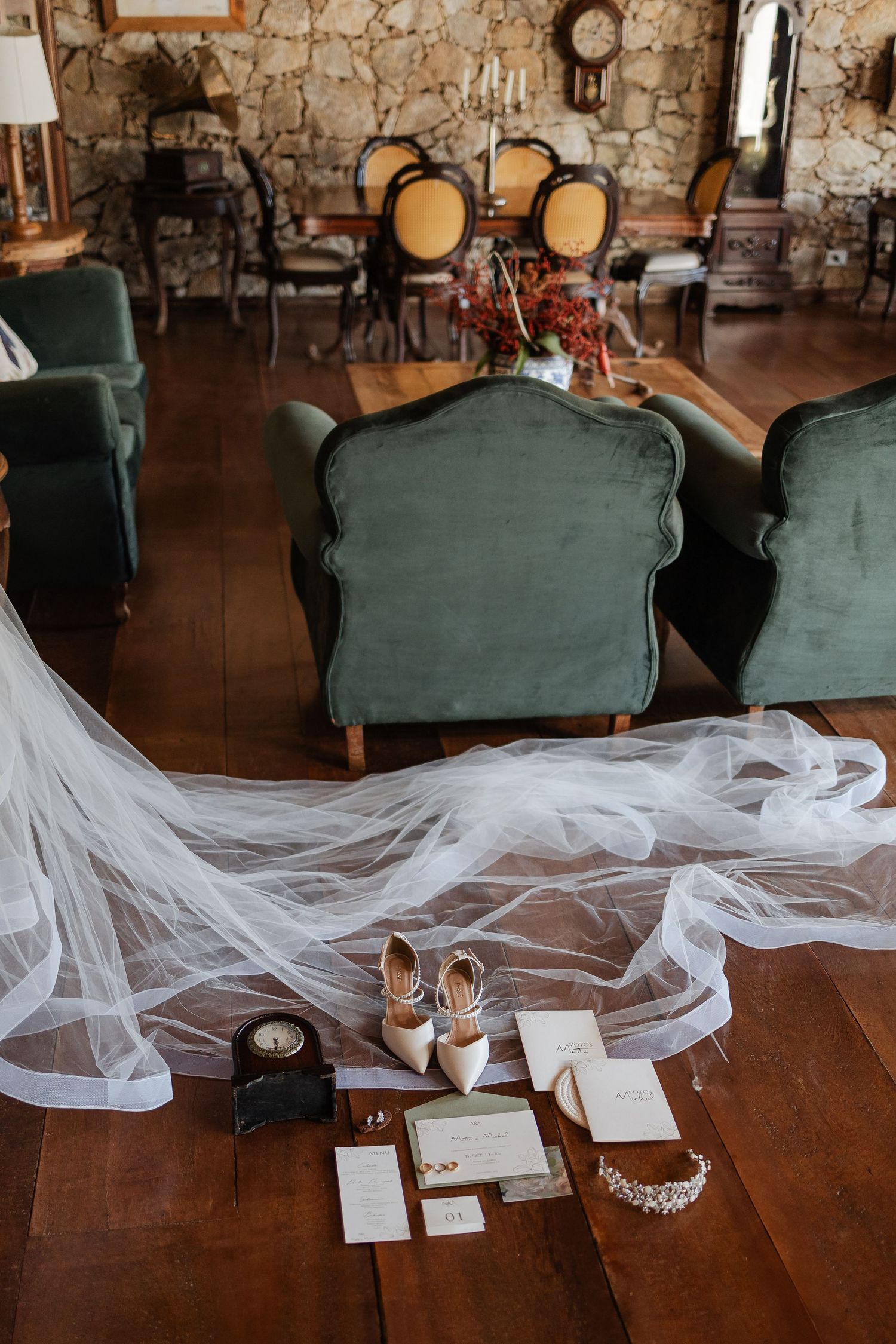 A long white wedding veil draped over green vintage chairs with wedding accessories and rings displayed on a wooden table.