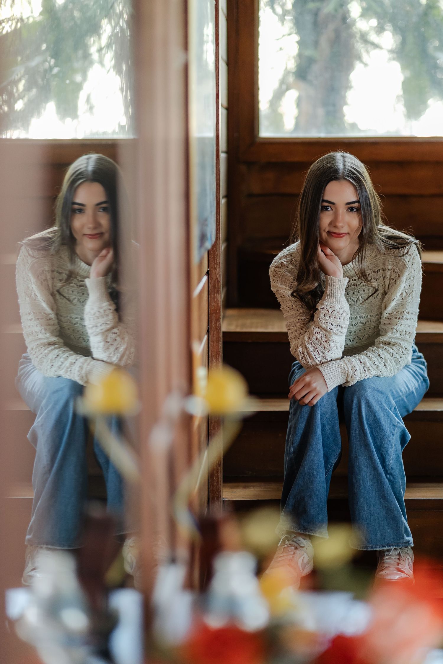 Mirror reflection of woman in white sweater and jeans sitting on wooden steps by window with flowers in foreground.