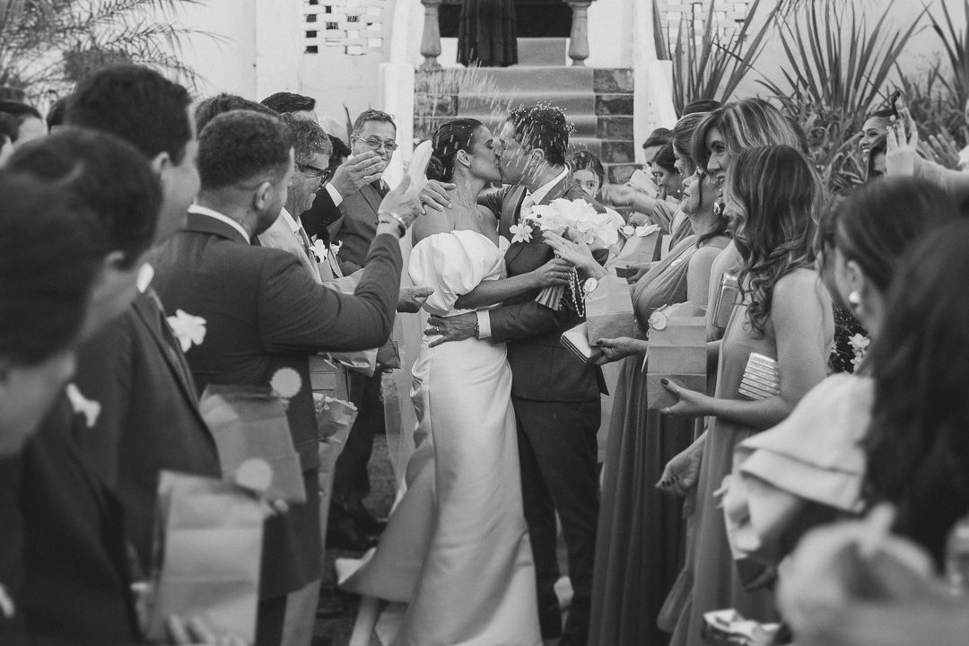 A vintage black and white photograph showing a crowded outdoor wedding reception with guests mingling among tropical plants.