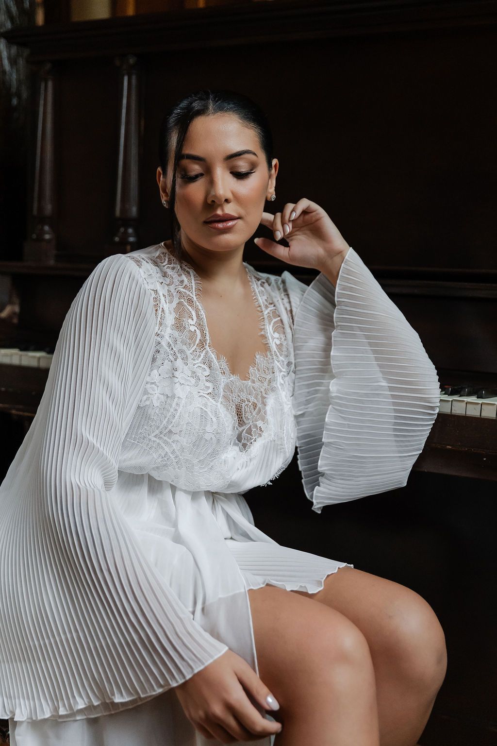 Fotógrafo de casamento em SP, Alfenas eElegant white lace and pleated robe with bell sleeves photographed next to a piano in moody lighting.