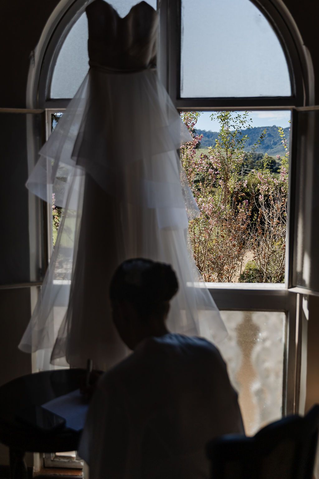 Cerimônia de casamento ao ar livreA white wedding dress hangs in an arched window while someone sits in silhouette against mountain scenery.