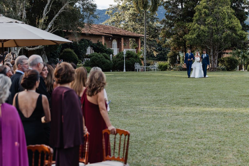 Wedding ceremony on a scenic lawn with guests seated on the left watching a special moment unfold near a brick house.
