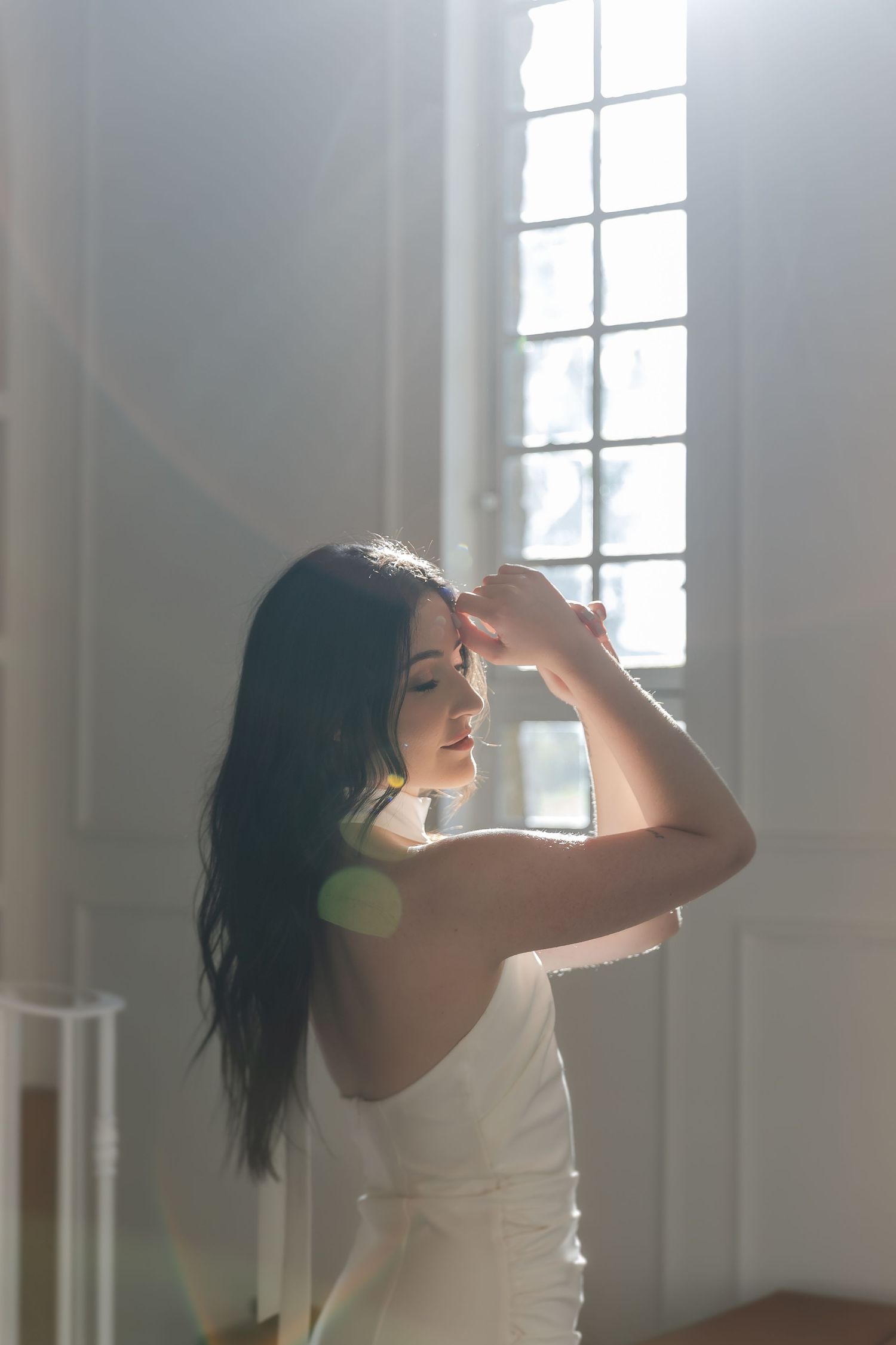 Dancer in white dress performs graceful movements in sunlit studio with large windows creating ethereal atmosphere.