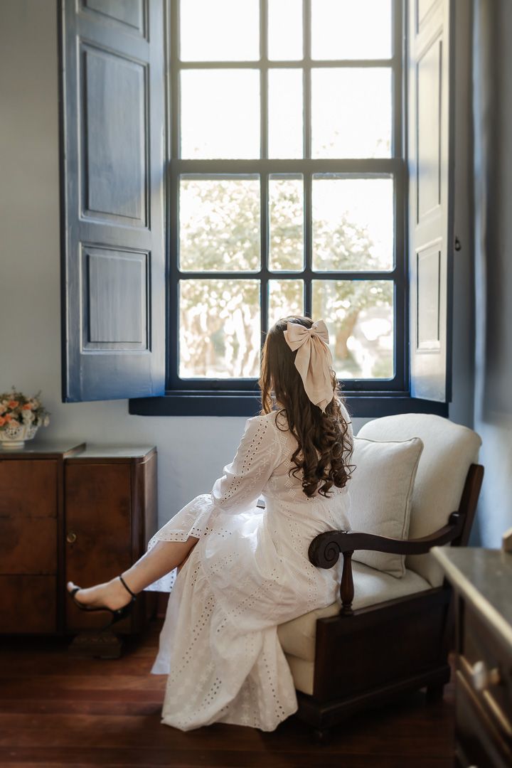 A person in a white lace dress sits near a window in a vintage chair, sunlight streaming through the room.