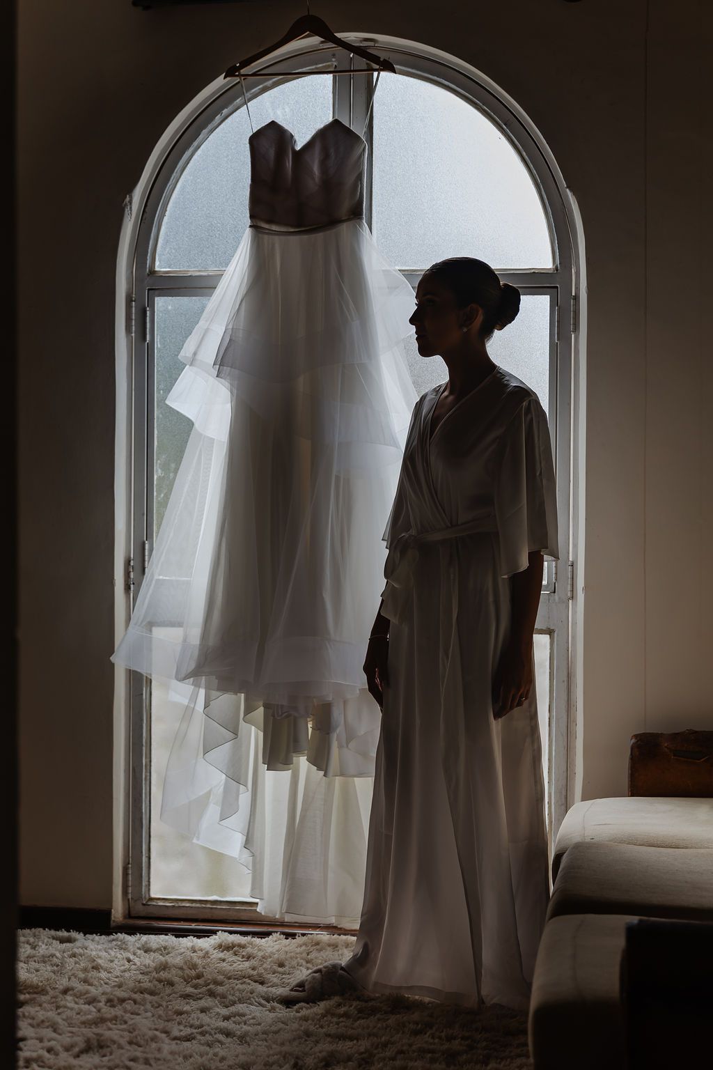 Cerimônia de casamento ao ar livre na FazenA silhouetted figure stands near an elegant white wedding gown hanging in an arched window frame.
