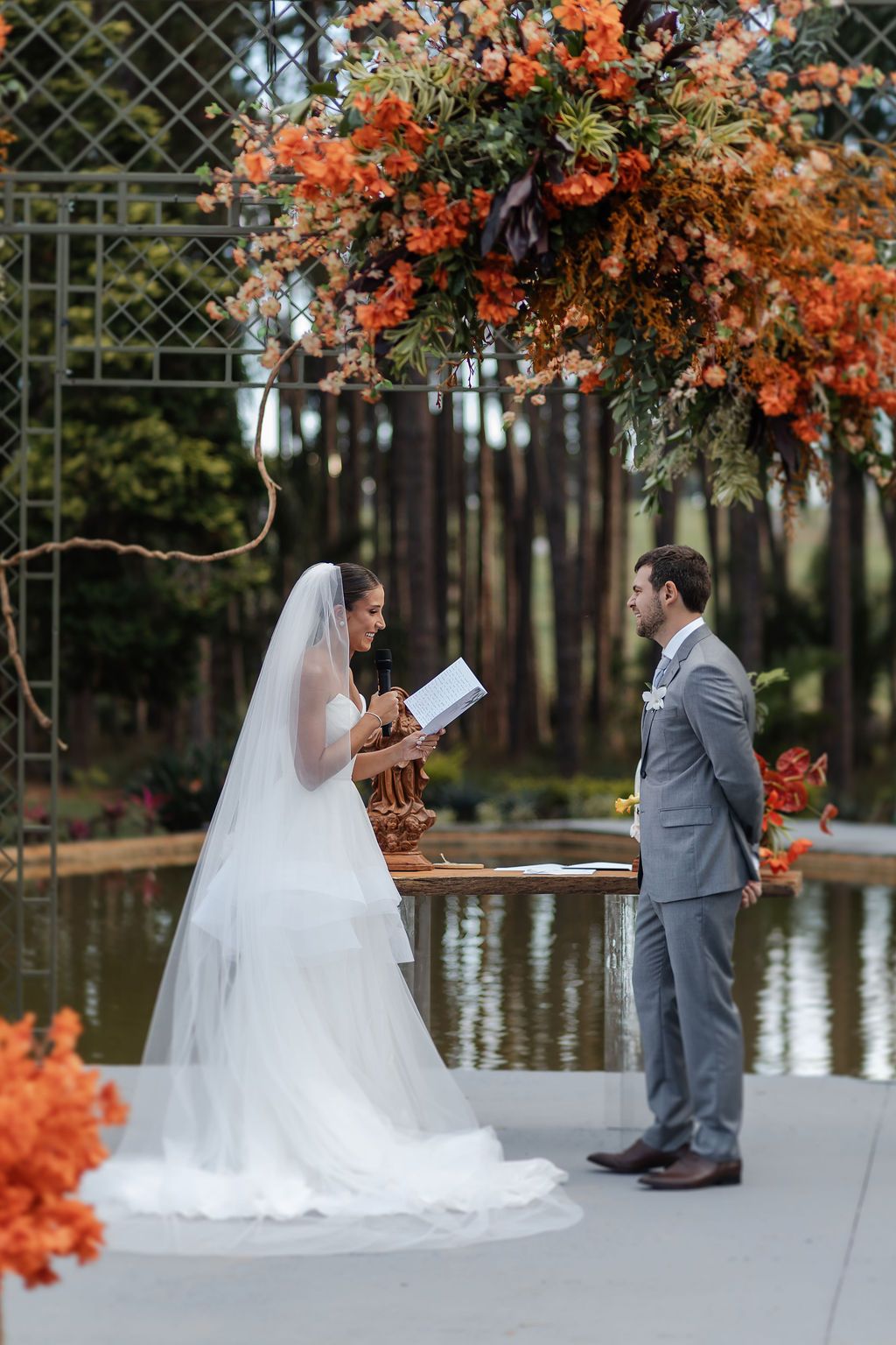 A wedding ceremony takes place on a dock with vibrant orange floral arrangements and a serene lake backdrop.
