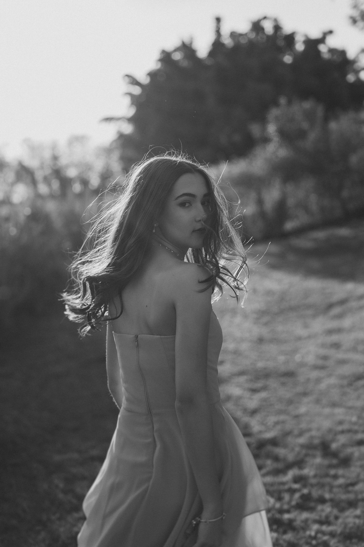 Woman in flowy white dress turns around in a backlit outdoor setting during golden hour creating a dreamy silhouette.