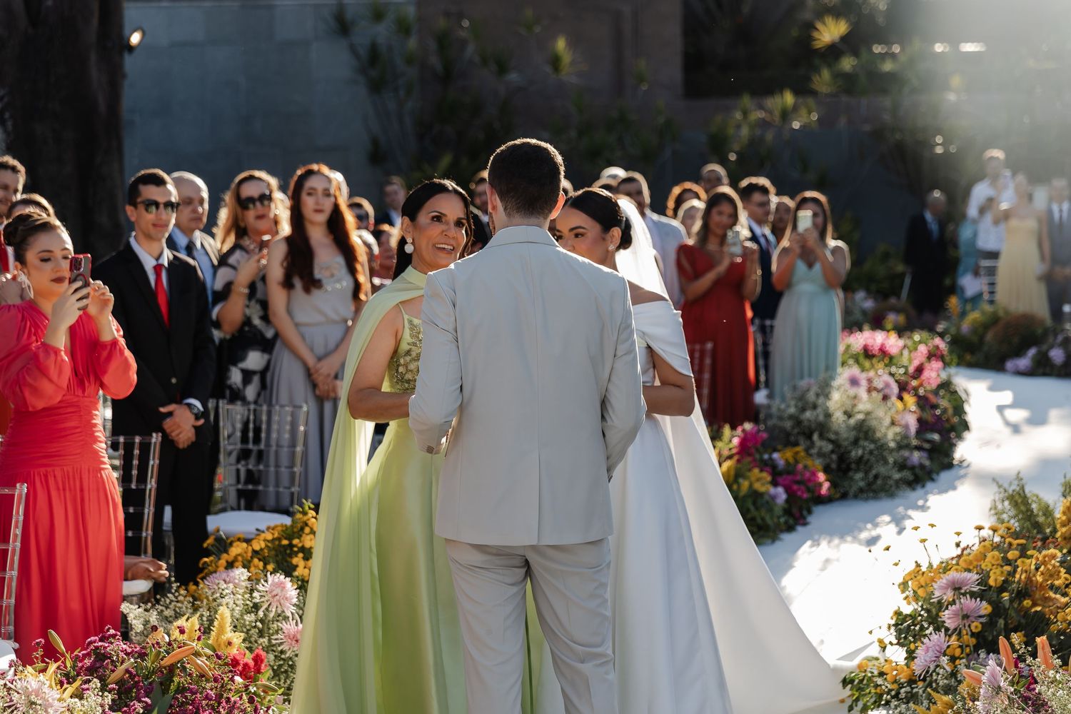 Guests in colorful dresses attend an outdoor garden wedding ceremony amid blooming flowers and sunlight.