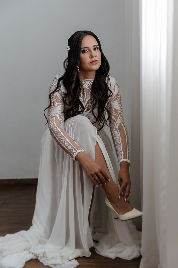An elegant long-sleeved white lace wedding dress with flowing skirt as the wearer adjusts their shoe near a window.