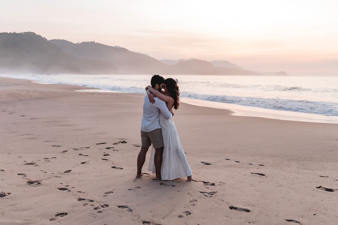A couple embraces on a misty beach at sunset with mountains and ocean waves in the background.
