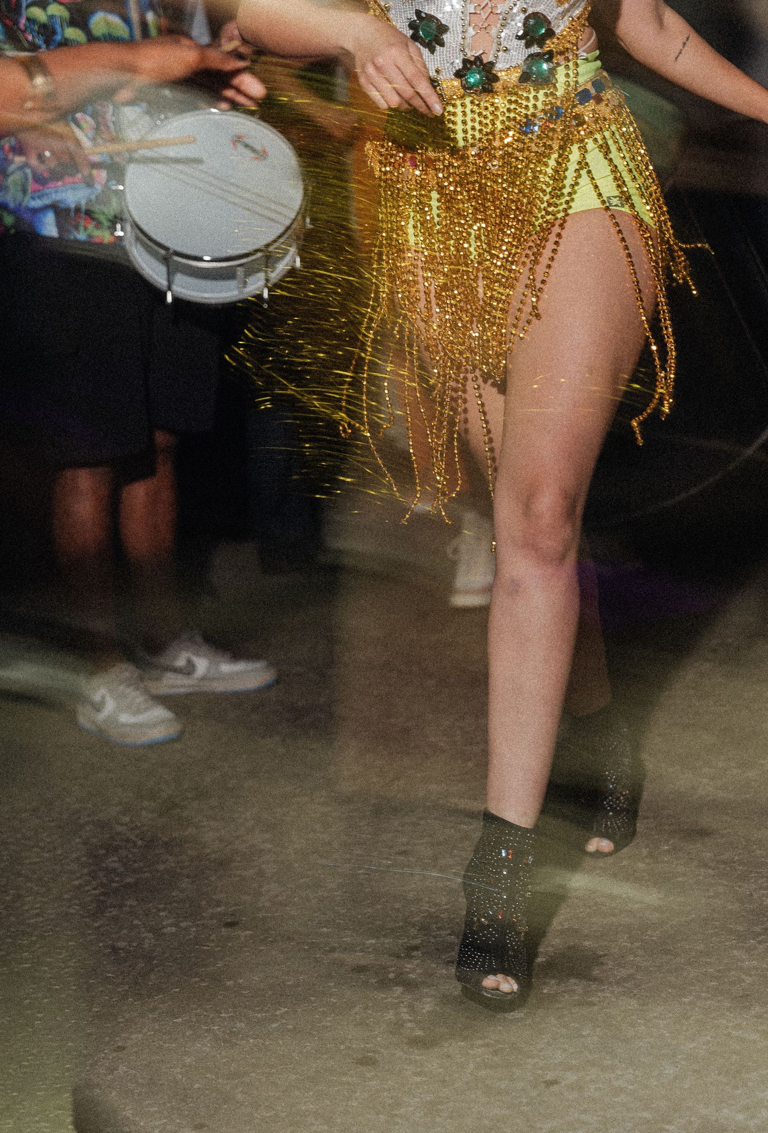 Dancers' legs in gold fringe skirts and various footwear move across a dark concrete floor at a party.