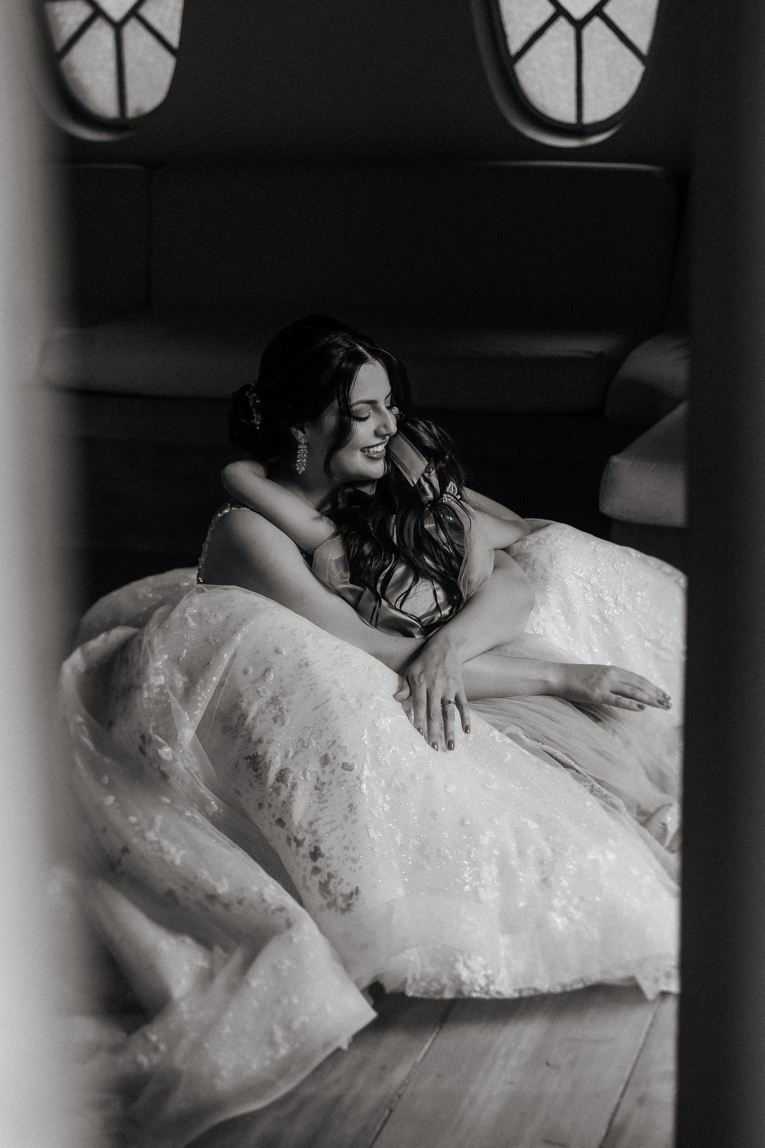A person in a white lace wedding dress reclines elegantly on a luxurious bed in a dramatic black and white photo.